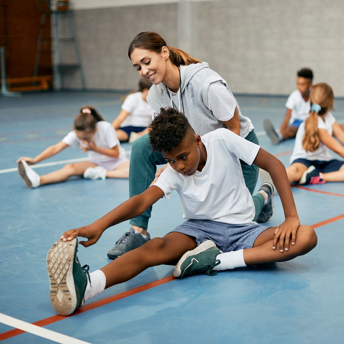 Black school boy stretching on the floor with help of PE teacher while warming up exercise class.