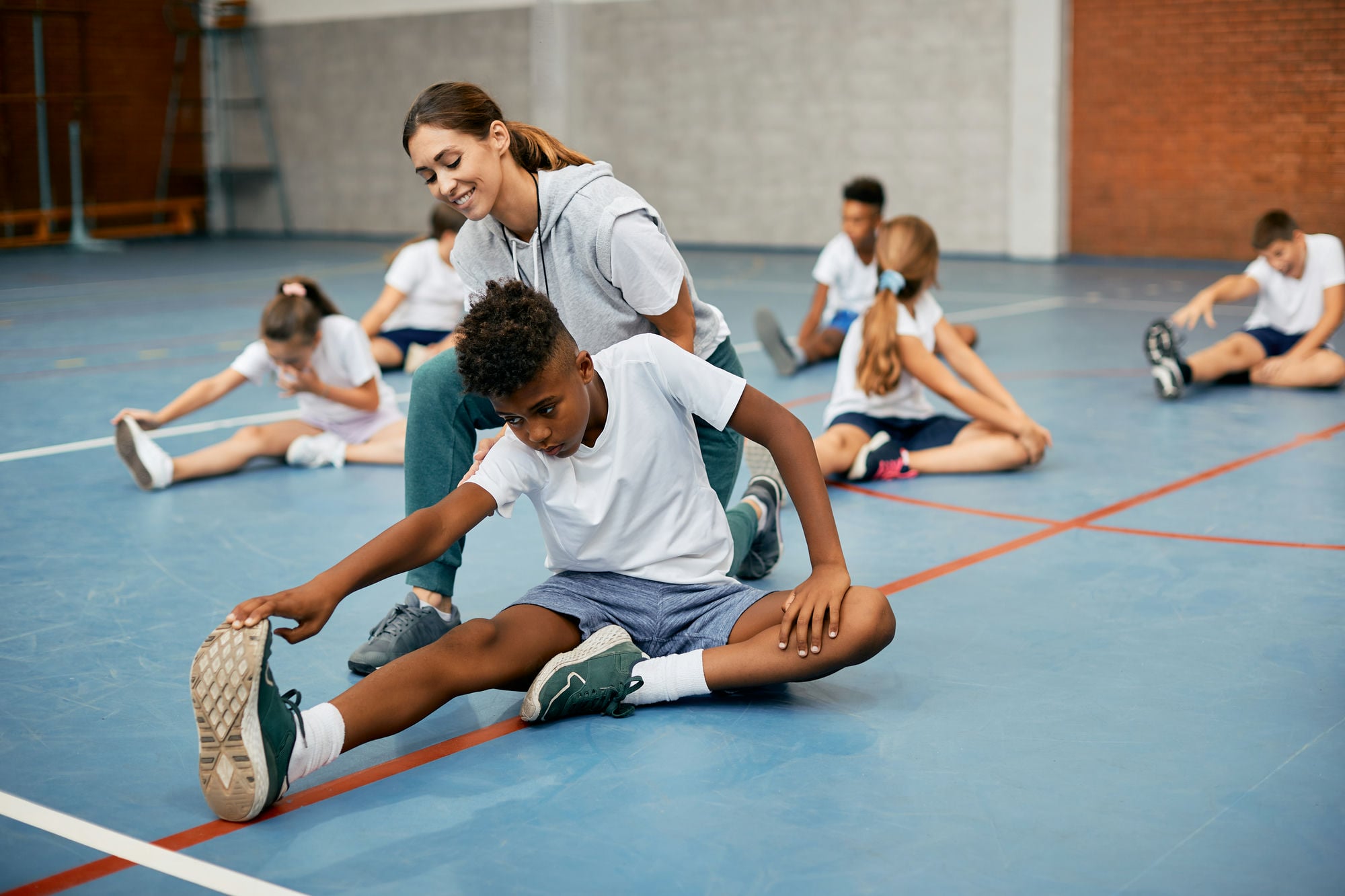 happy-female-sports-teacher-assisting-african-amer-2024-12-13-18-36-29-utc_min Black school boy stretching on the floor with help of PE teacher while warming up exercise class.