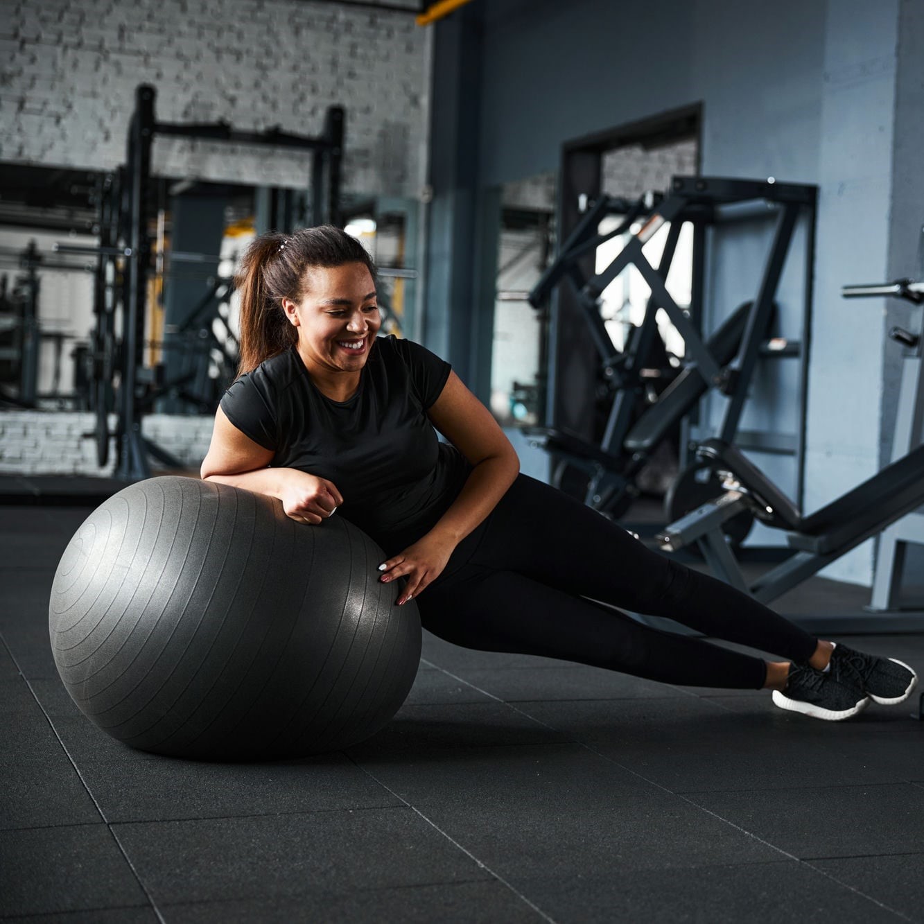 Cheerful pretty woman is using fitball for balancing on it in side plank position in gym